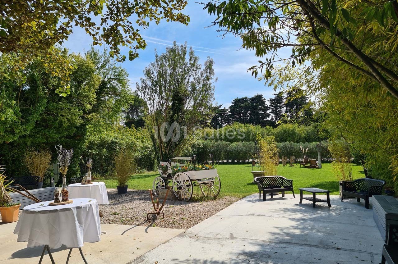 Jardin verdoyant avec pelouse, arbres, wagon décoratif et tables dressées sur une terrasse ensoleillée.