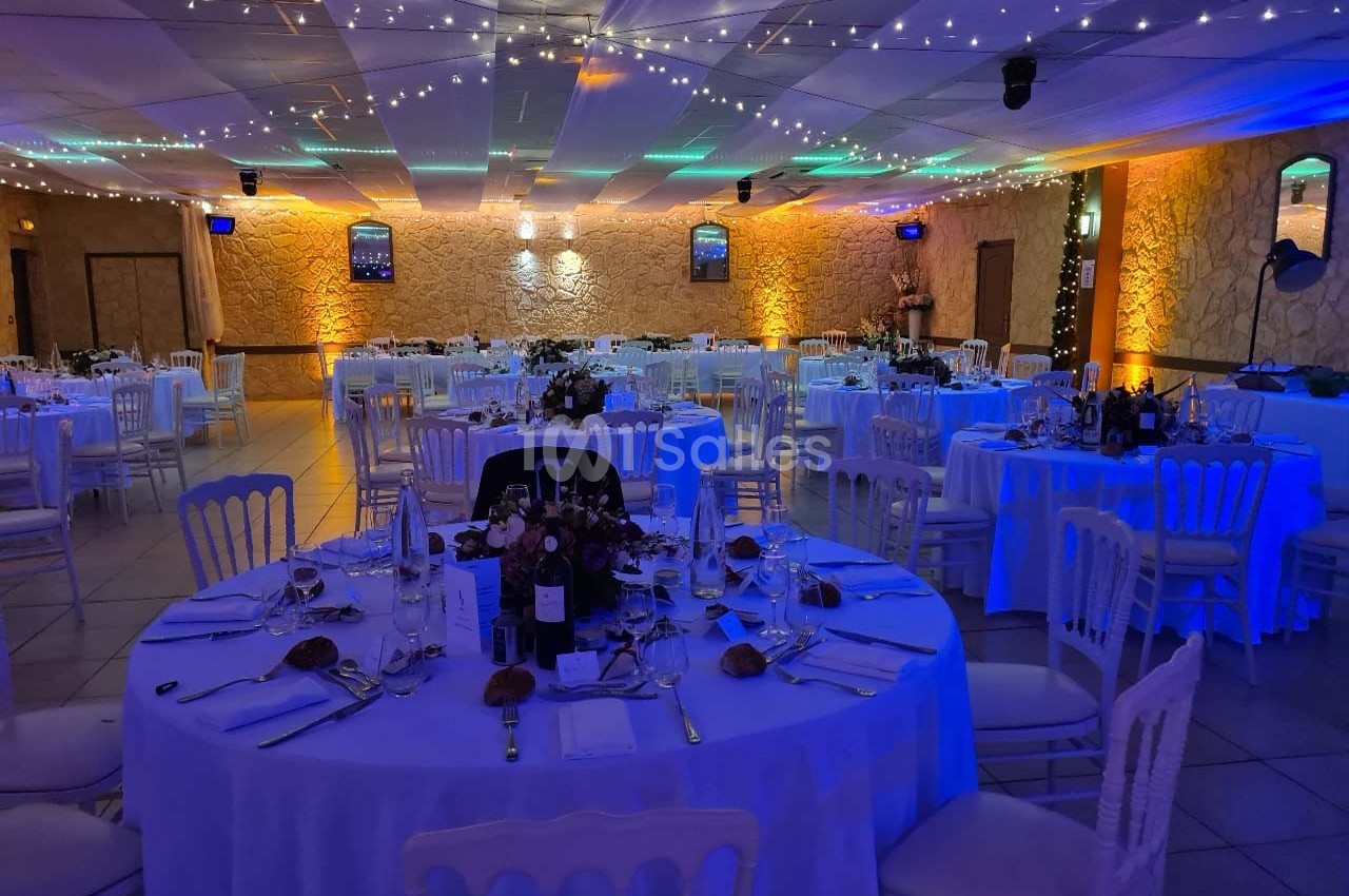 Salle de réception décorée avec des tables rondes dressées, éclairage tamisé et guirlandes lumineuses au plafond.