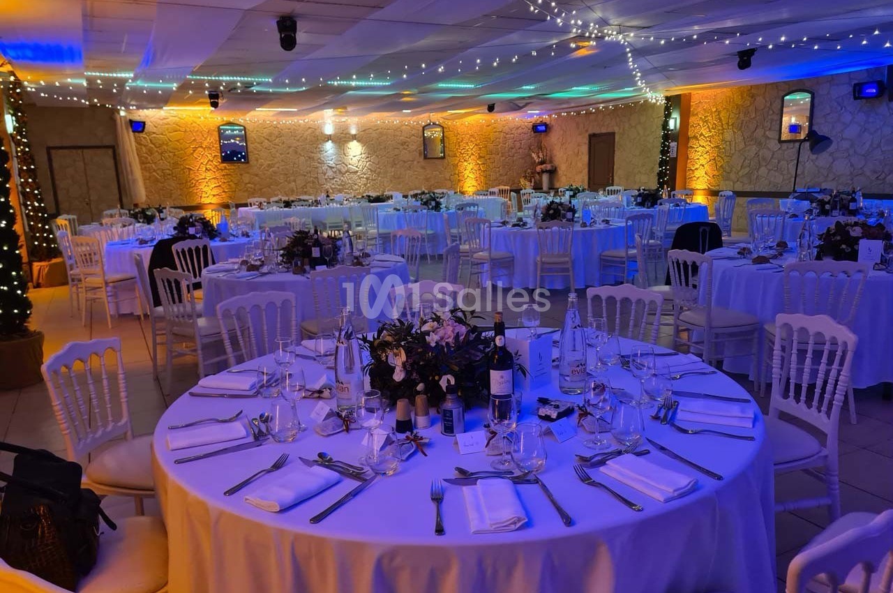 Salle de réception décorée avec des tables rondes dressées, éclairage tamisé et guirlandes lumineuses au plafond.