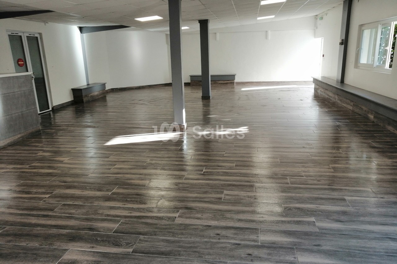 Salle vide avec un sol en parquet sombre, murs blancs, fenêtres et piliers gris sous un plafond à dalles blanches.
