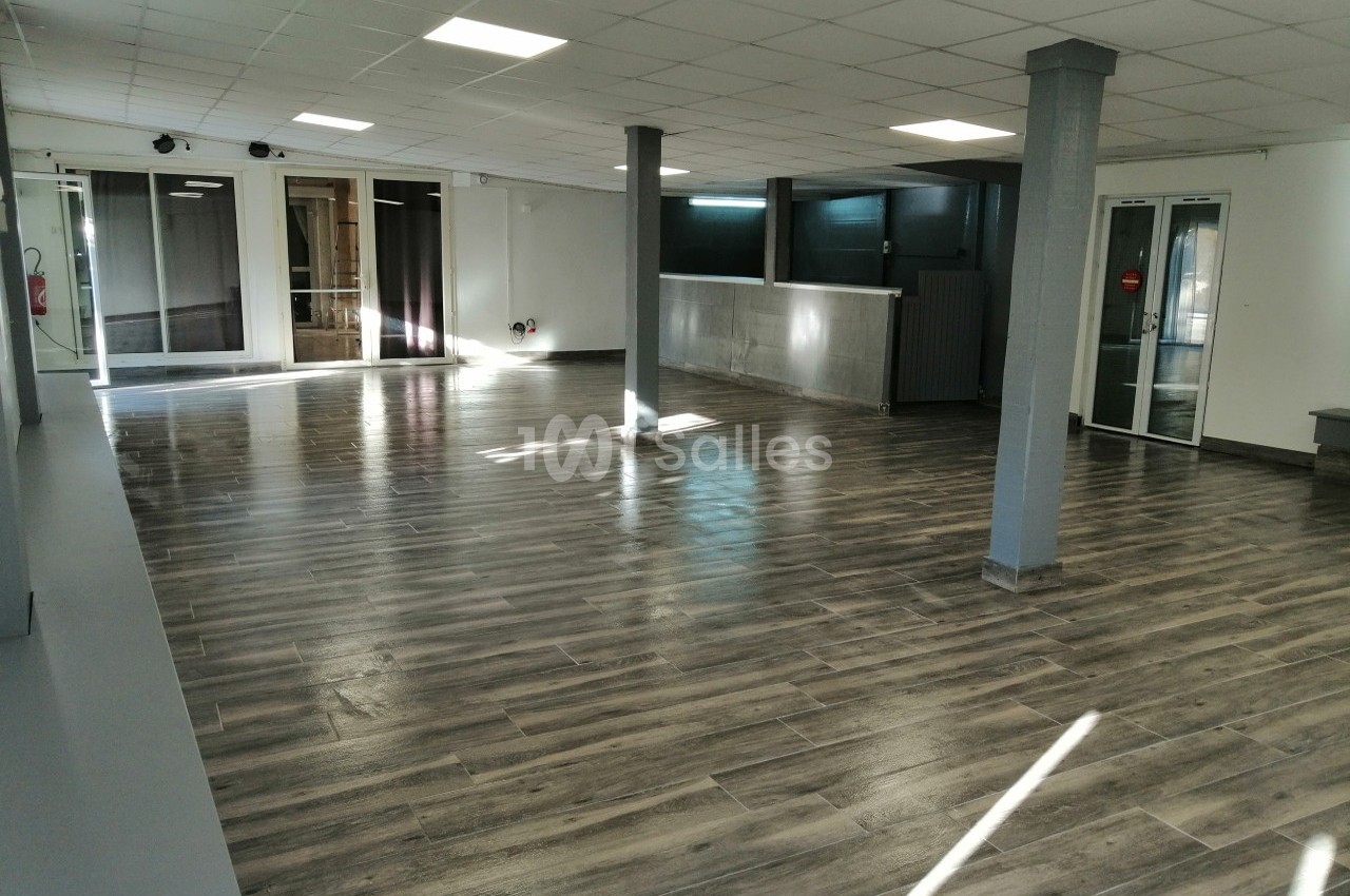 Salle vide avec sol en parquet gris, piliers centraux, murs clairs et grandes baies vitrées laissant entrer la lumière.