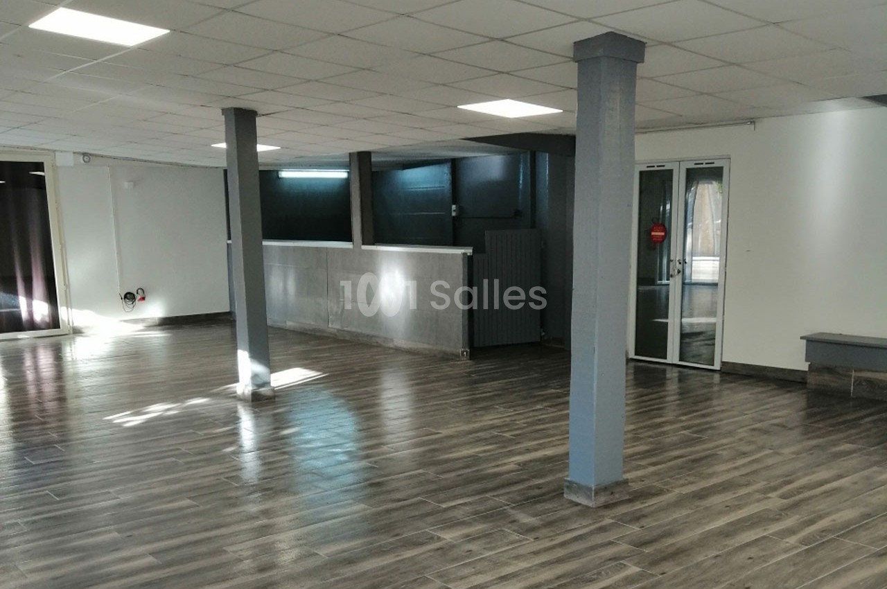 Salle vide avec sol en parquet foncé, murs clairs, plafond à dalles blanches et grandes baies vitrées.