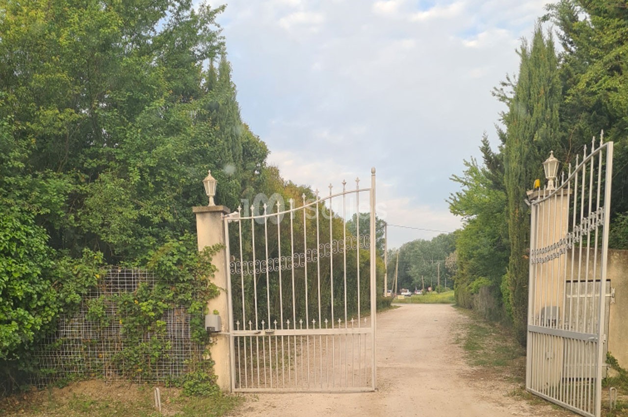 Portail métallique blanc ouvert donnant sur un chemin de terre bordé de végétation et d'arbres.