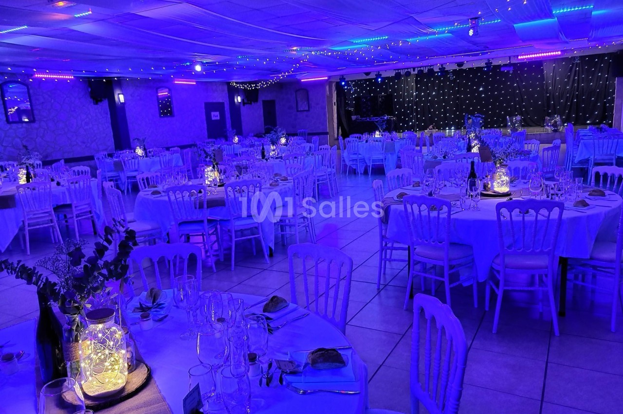 Salle de réception décorée avec des tables rondes dressées, éclairage bleu et guirlandes lumineuses au plafond.
