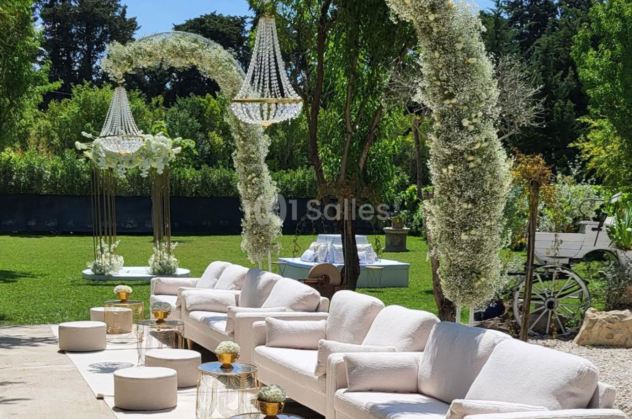 Espace extérieur décoré pour un événement avec canapés blancs, arches fleuries et lustres suspendus.