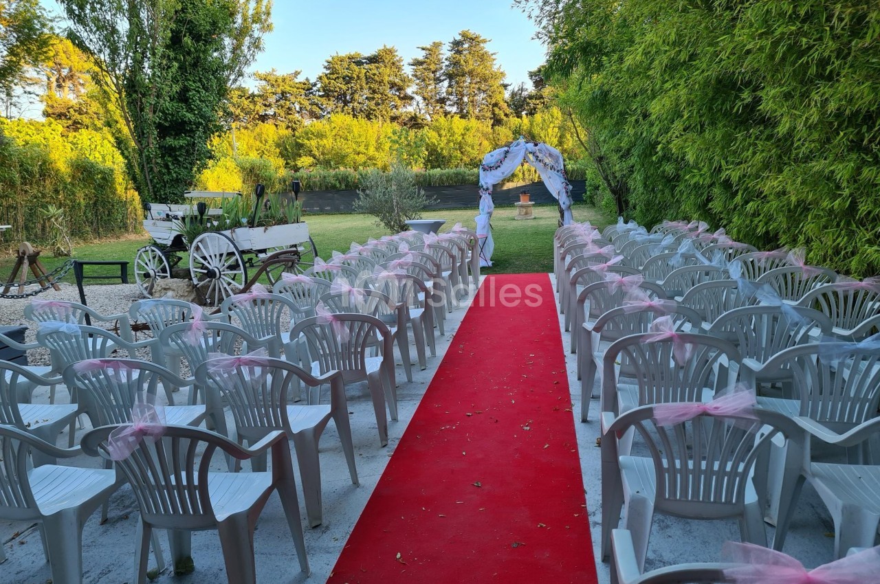 Allée centrale avec tapis rouge entourée de chaises blanches décorées, menant à une arche fleurie dans un jardin.
