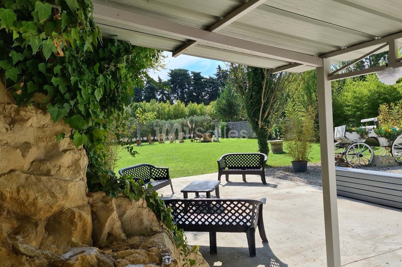 Terrasse couverte avec mobilier en fer forgé, vue sur un jardin verdoyant et des éléments décoratifs extérieurs.