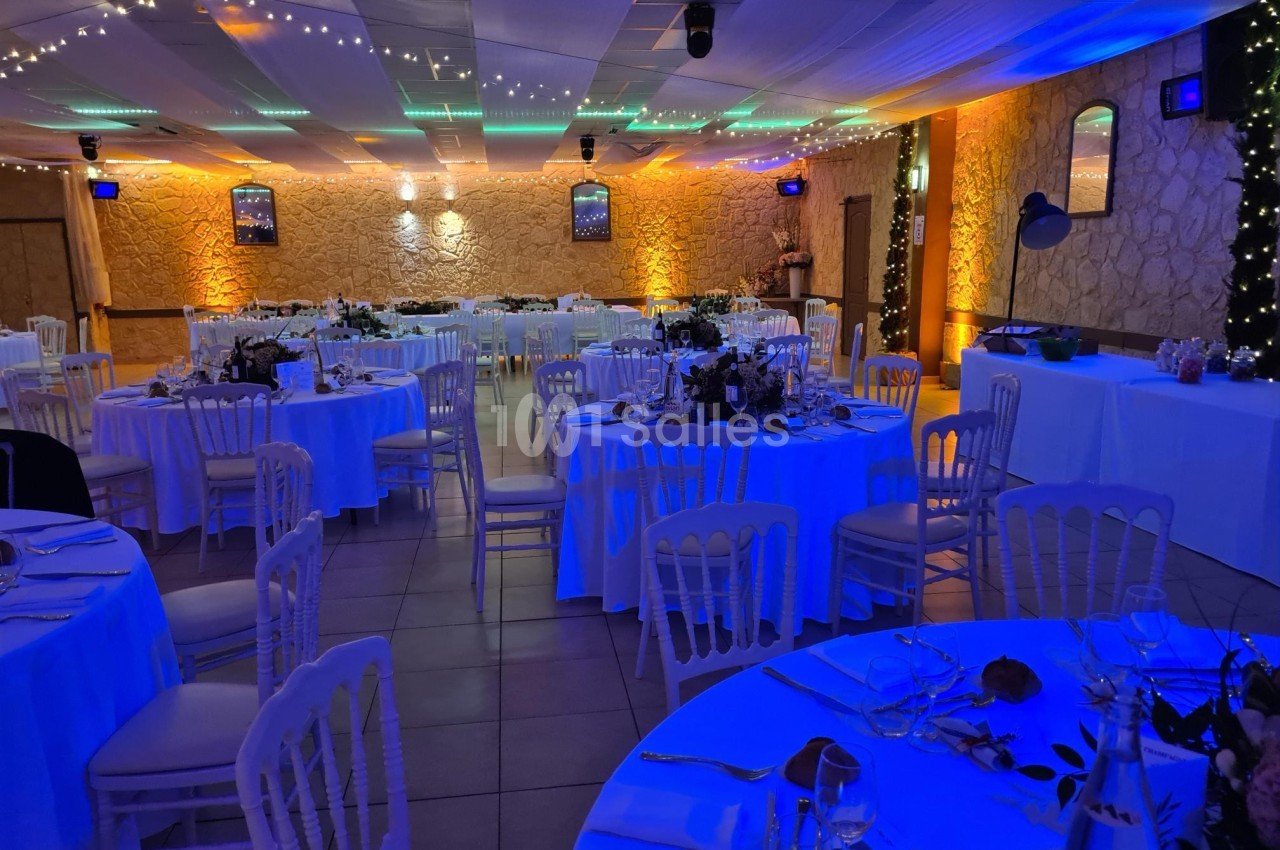 Salle de réception décorée avec des tables rondes dressées, éclairage chaleureux et guirlandes lumineuses au plafond.