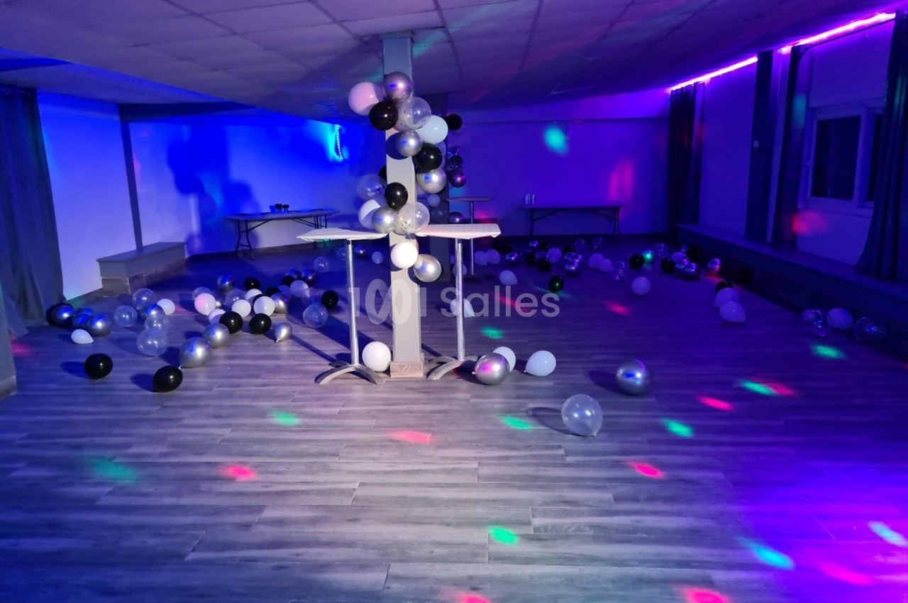 Salle vide décorée de ballons noirs, blancs et argentés, éclairée par des lumières colorées sur un sol en parquet.