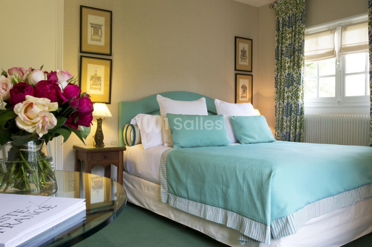 Chambre lumineuse avec lit double, couvre-lit turquoise, fleurs sur une table et fenêtre encadrée de rideaux à motifs.