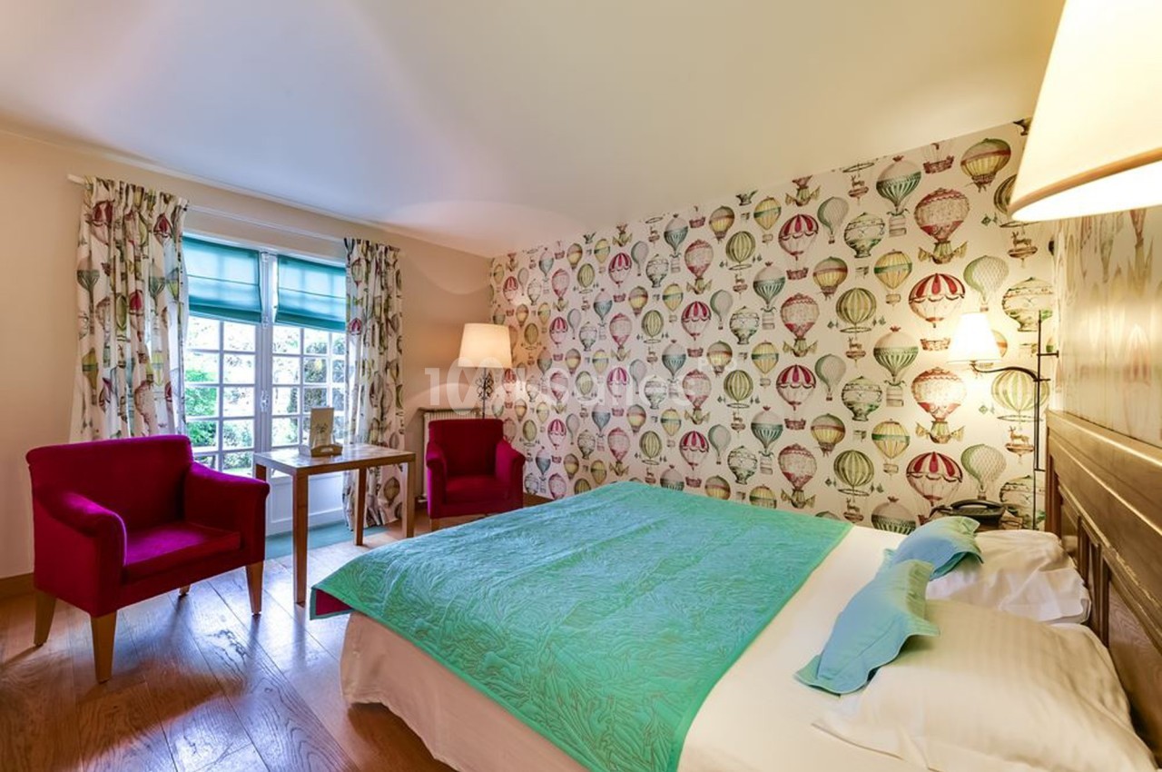 Chambre lumineuse avec lit double, couvre-lit vert, fauteuils rouges, lampe et fenêtre donnant sur un jardin.
