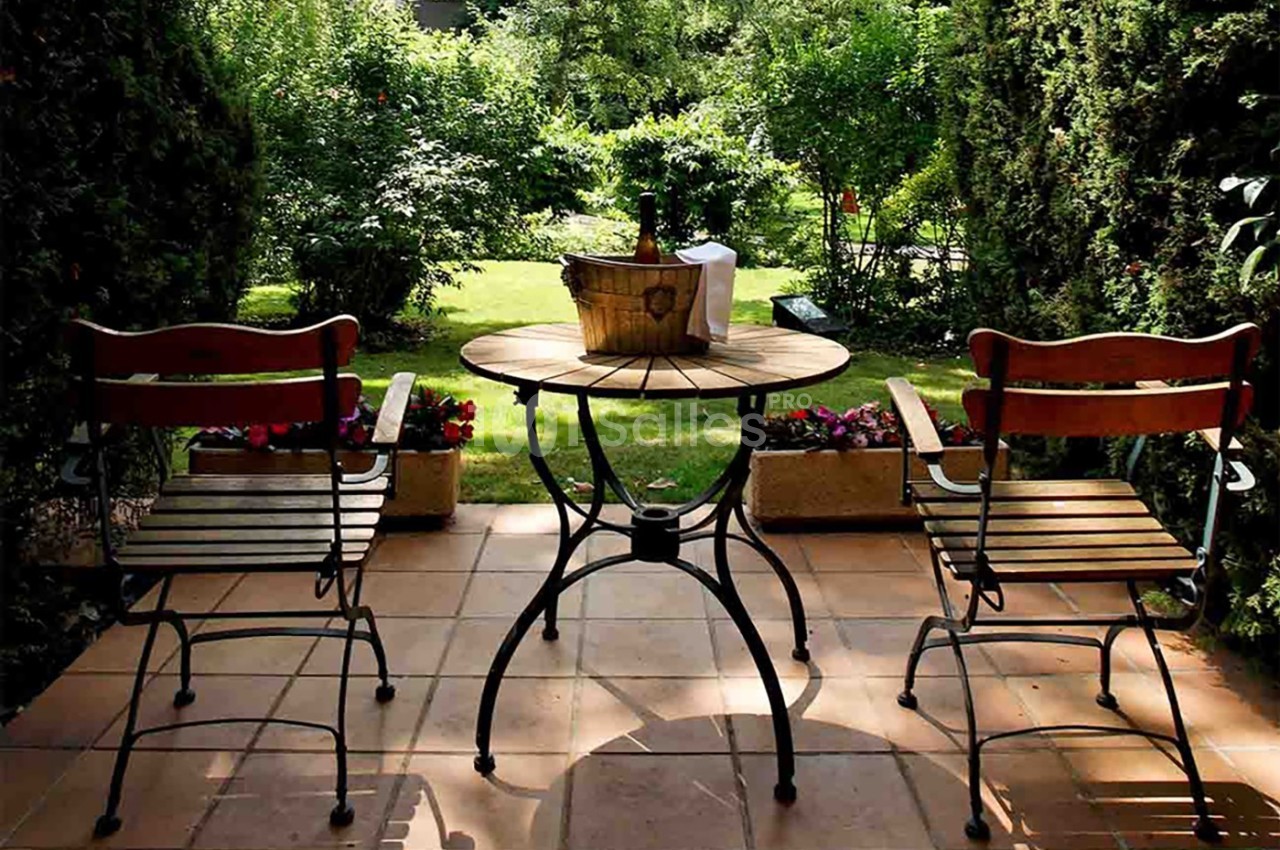 Deux chaises en bois et une table ronde avec un seau à glace sur une terrasse donnant sur un jardin verdoyant.