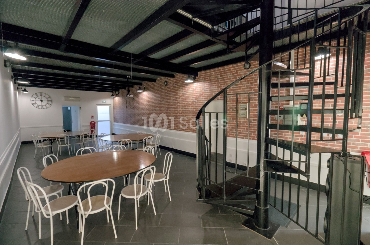 Salle avec tables rondes, chaises blanches, mur en briques, escalier en colimaçon et plafond industriel.