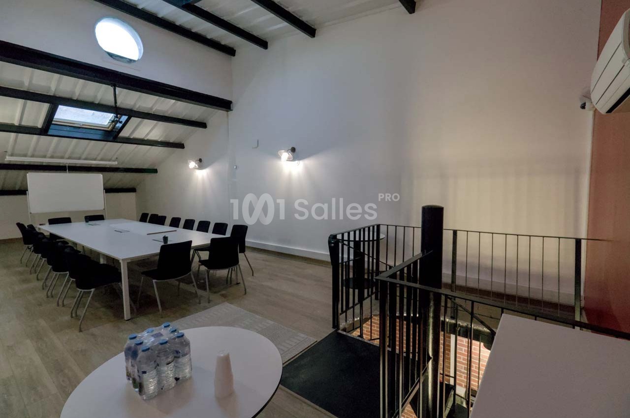 Salle de réunion lumineuse avec une grande table, des chaises noires, un tableau blanc et un escalier en premier plan.