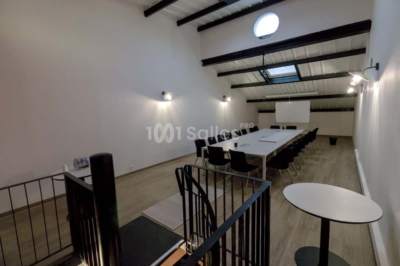 Salle de réunion lumineuse avec une grande table, des chaises noires, un tableau blanc et un plafond incliné.