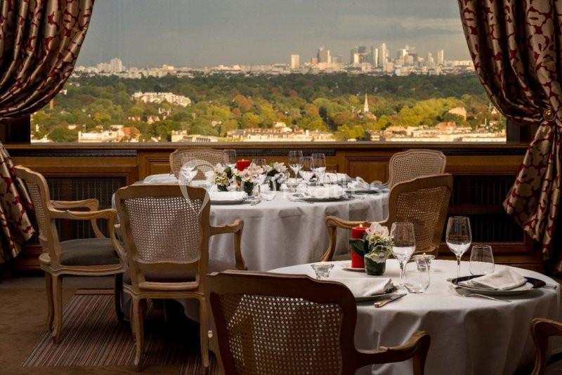 Salle de restaurant élégante avec tables dressées, vue panoramique sur une ville et un paysage verdoyant en arrière-plan.