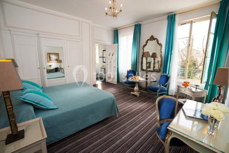 Chambre lumineuse avec lit double, fauteuils bleus, table, miroir doré et grandes fenêtres donnant sur l'extérieur.
