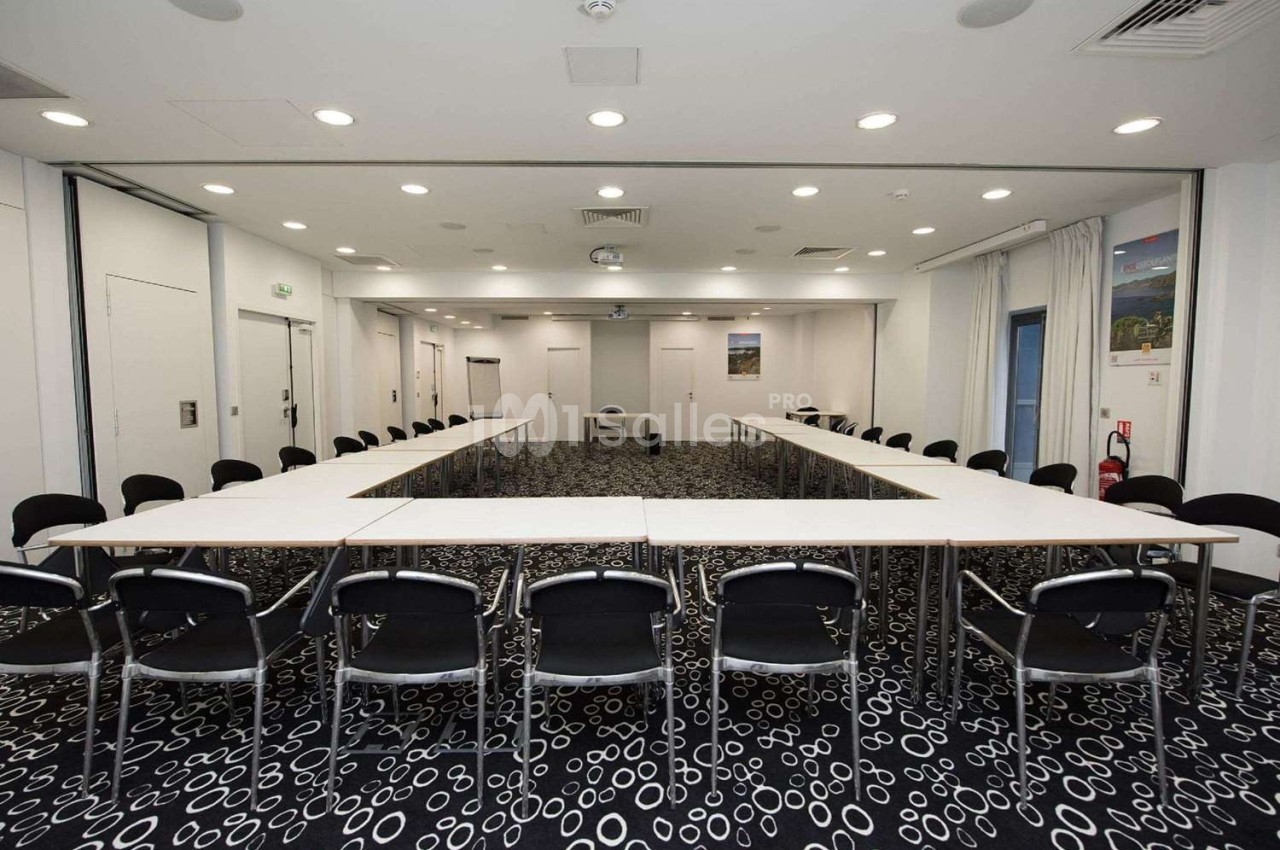 Salle de réunion avec des tables disposées en U, des chaises noires et un éclairage lumineux.