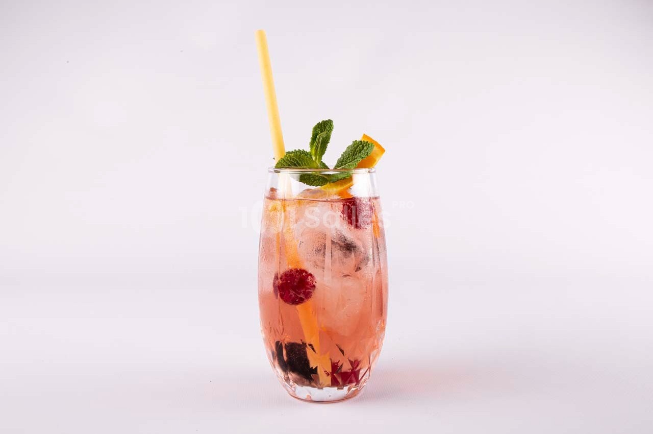 Verre de cocktail rose garni de glaçons, framboises, mûres, tranche d'orange, feuilles de menthe et une paille jaune.