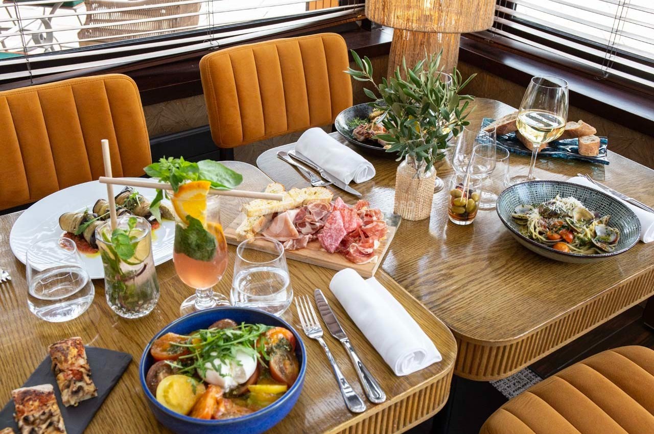 Table dressée avec des plats variés, boissons, couverts et décoration végétale dans un cadre chaleureux.