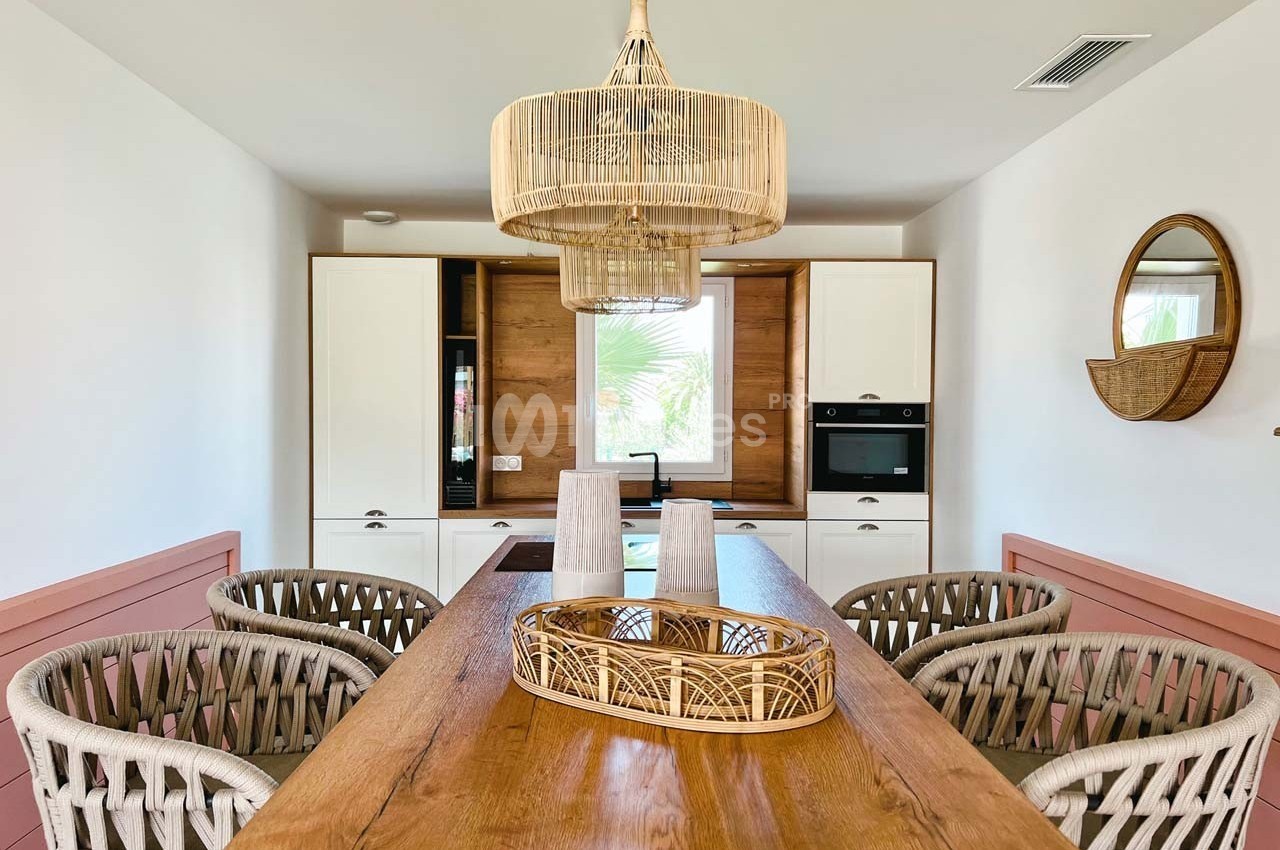 Salle à manger lumineuse avec table en bois, chaises tressées, suspension en rotin et cuisine équipée en arrière-plan.