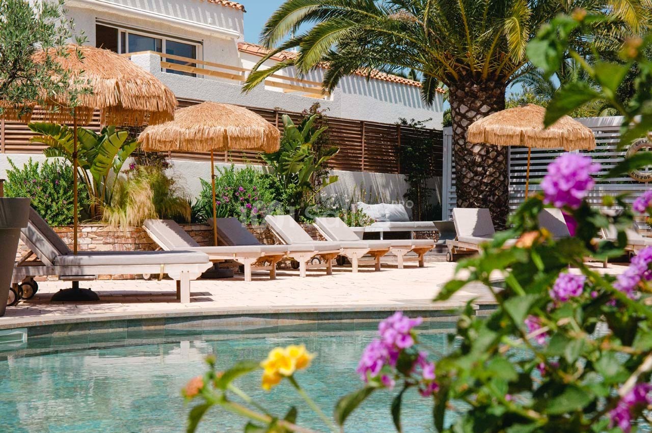 Terrasse avec piscine, transats en bois, parasols en paille, végétation tropicale et maison en arrière-plan.