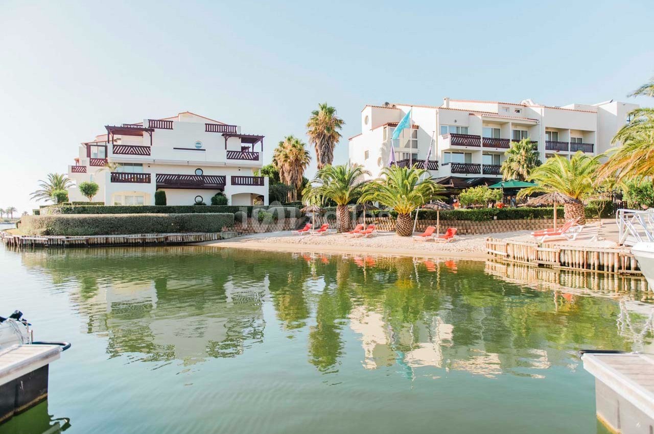 Vue sur des bâtiments résidentiels blancs avec balcons, entourés de palmiers, au bord d'un plan d'eau calme.
