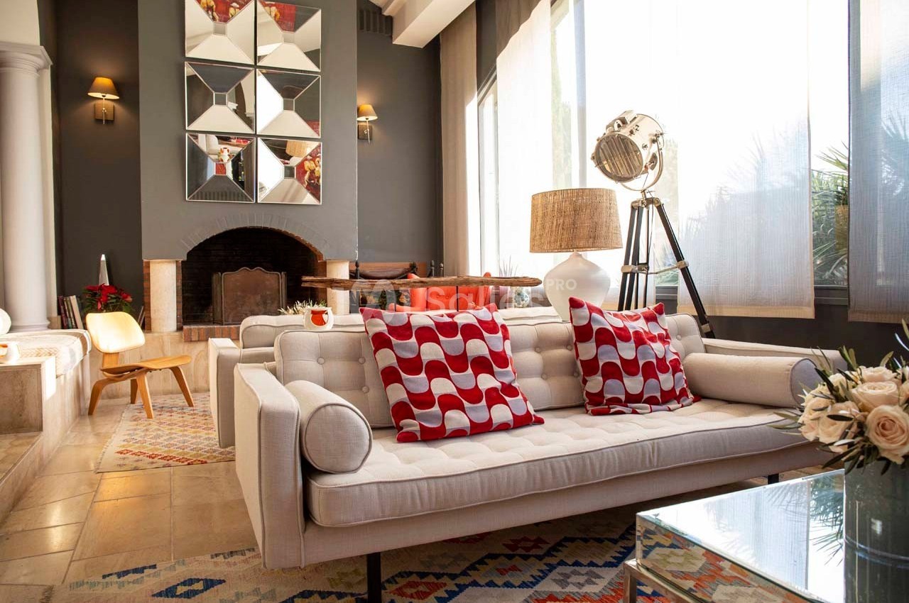 Salon moderne avec canapé blanc, coussins rouges à motifs, cheminée, lampadaire et grande fenêtre laissant entrer la lumière.