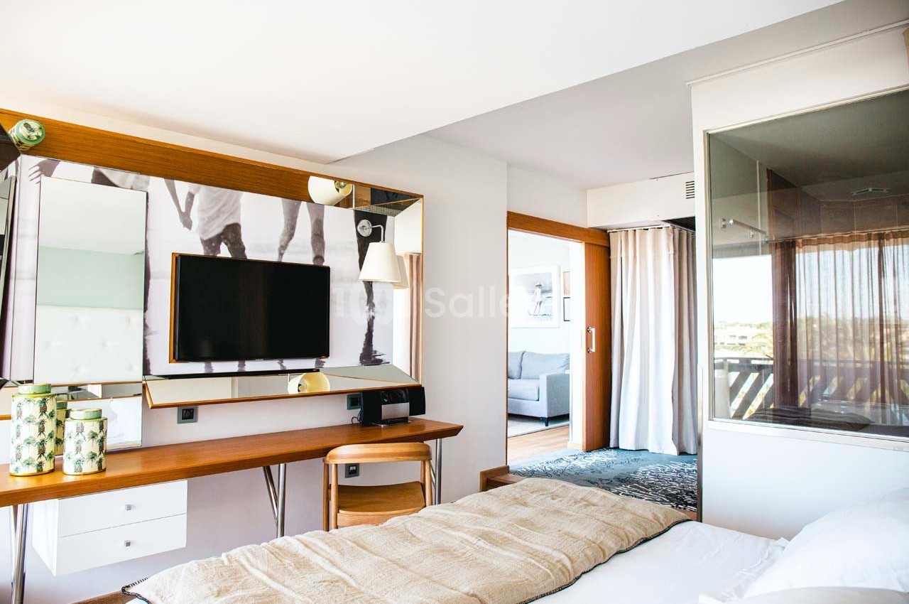 Chambre lumineuse avec lit, bureau en bois, télévision murale et vue sur un salon à travers une porte ouverte.