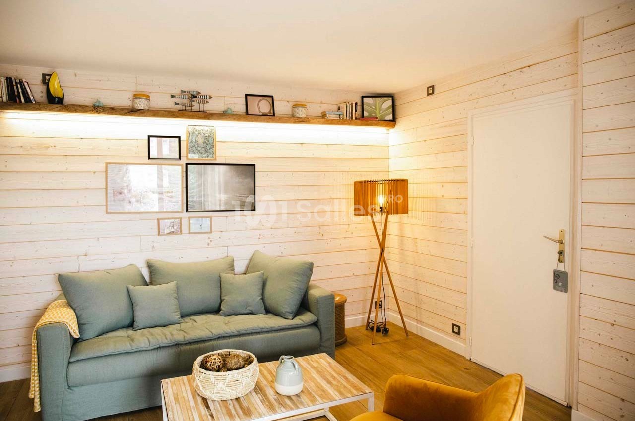 Salon lumineux avec murs en bois clair, canapé bleu, fauteuil jaune, lampe sur pied et étagère décorative.