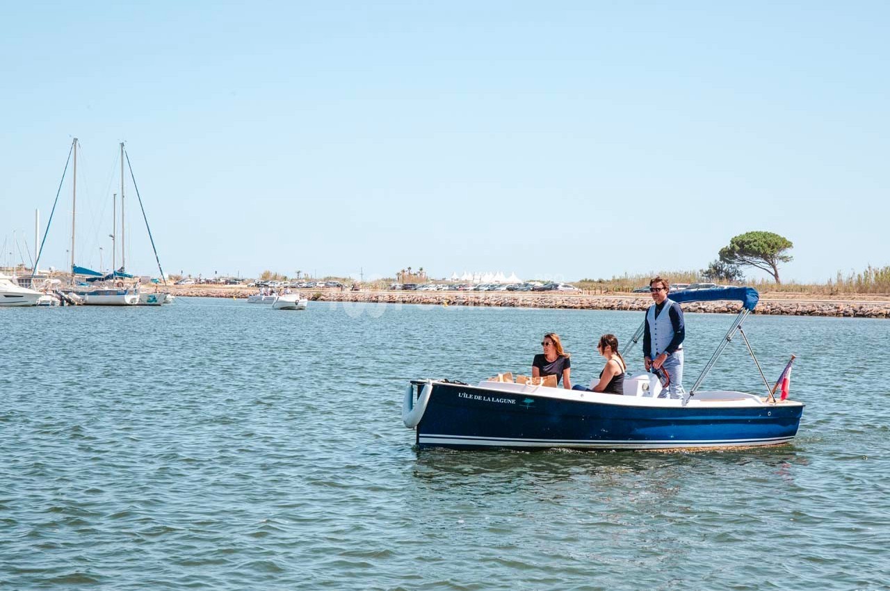 Un petit bateau à moteur navigue sur une eau calme avec trois personnes à bord, un port visible en arrière-plan.