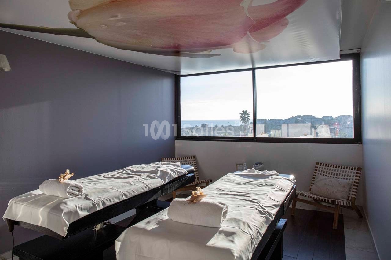Salle de massage lumineuse avec deux tables de soins, vue sur l'extérieur et plafond décoré d'une fleur.