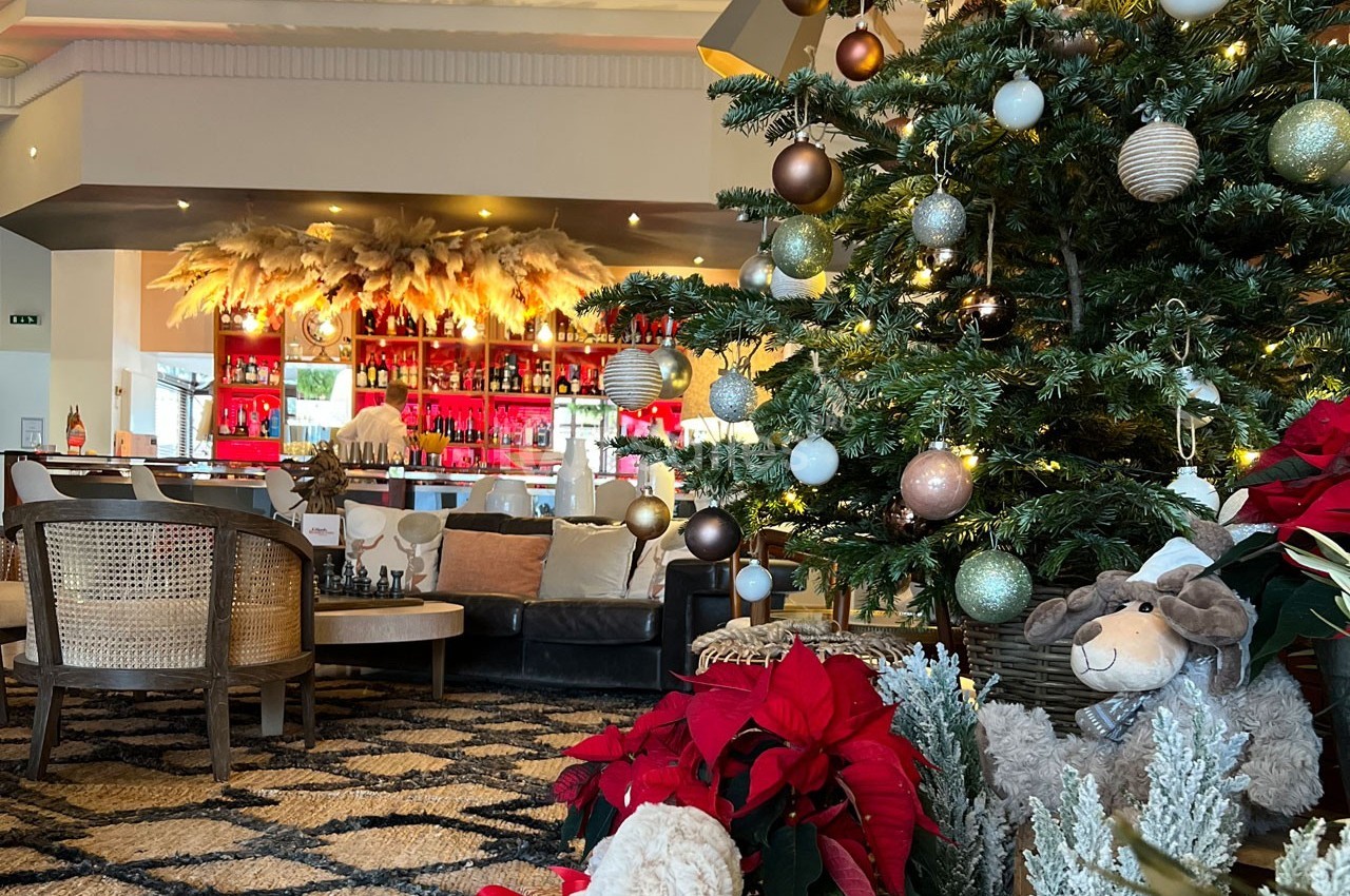 Sapin de Noël décoré avec des boules et guirlandes, entouré de poinsettias, dans un salon avec un bar en arrière-plan.