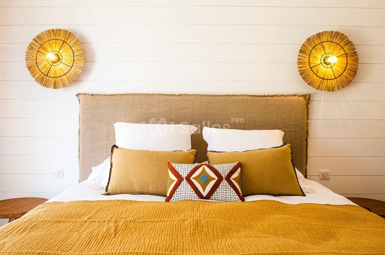 Lit double avec tête de lit en tissu beige, coussins décoratifs et luminaires muraux en rotin sur fond de mur blanc.