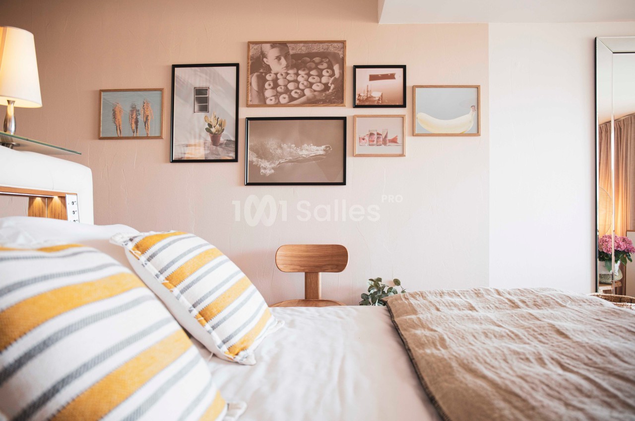 Mur décoré de cadres variés dans une chambre lumineuse avec lit, coussins rayés et chaise en bois.