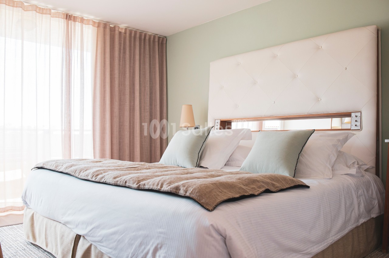 Lit double avec tête de lit capitonnée, draps blancs, coussins beiges, dans une chambre lumineuse aux rideaux roses.
