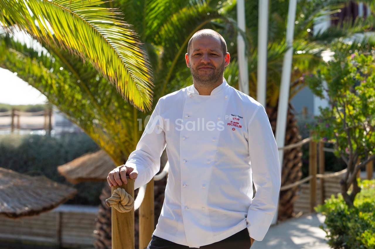 Un chef en veste blanche pose devant des palmiers et une terrasse ensoleillée.