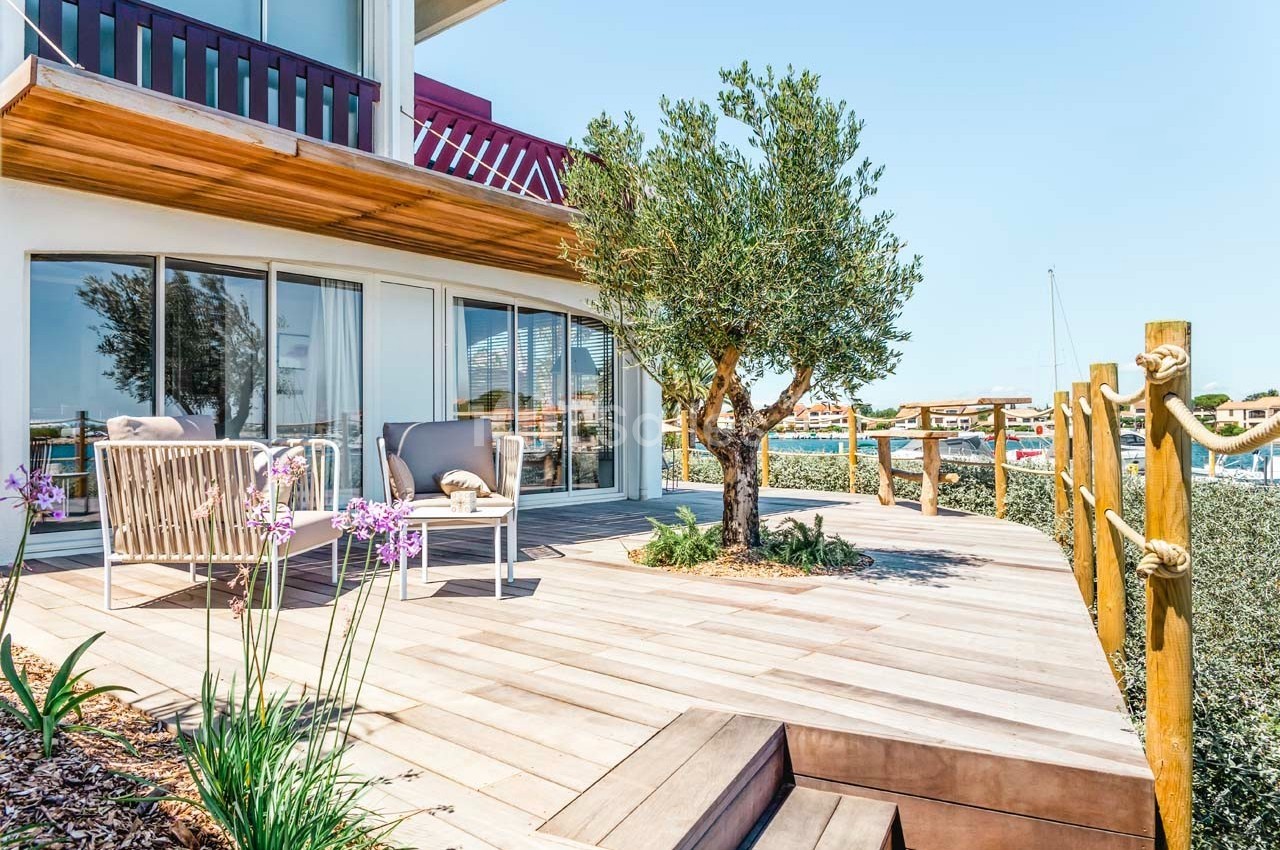 Terrasse en bois avec fauteuils, table basse, arbre et vue sur un port de plaisance en arrière-plan.