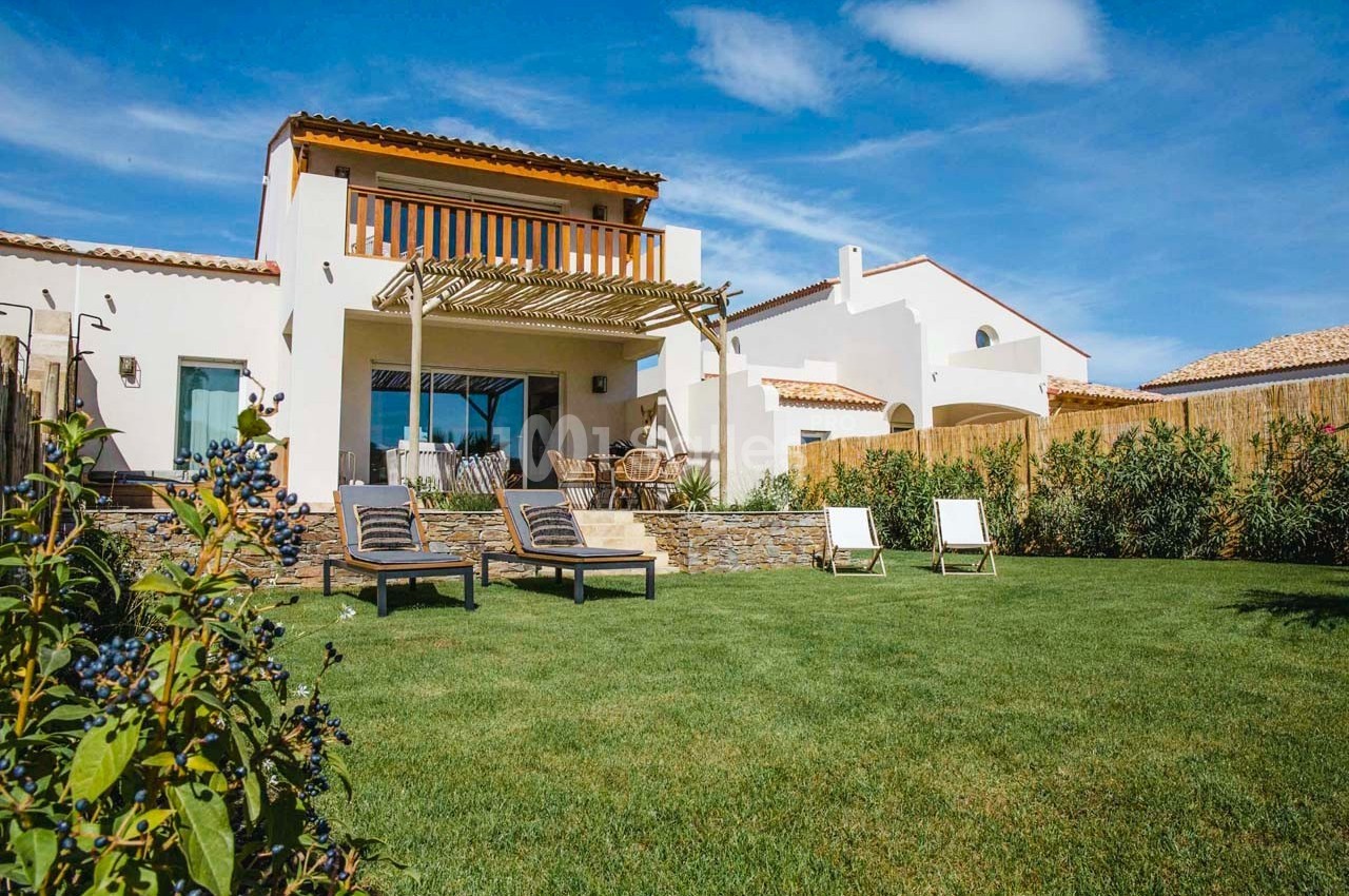 Maison moderne avec terrasse en bois, chaises longues et jardin verdoyant sous un ciel bleu.