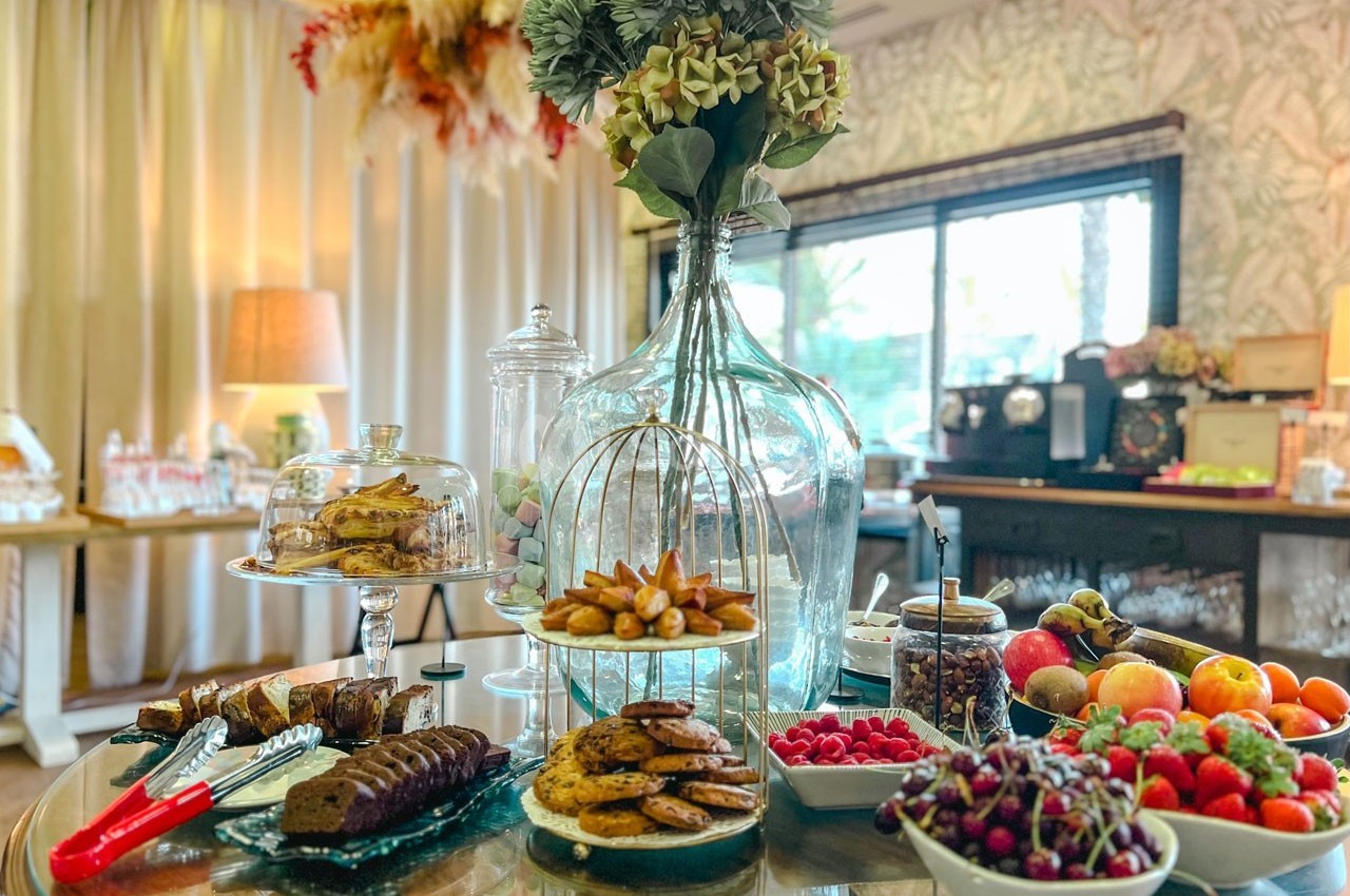 Table garnie de desserts variés, fruits frais, biscuits et pâtisseries, dans une salle lumineuse et décorée.