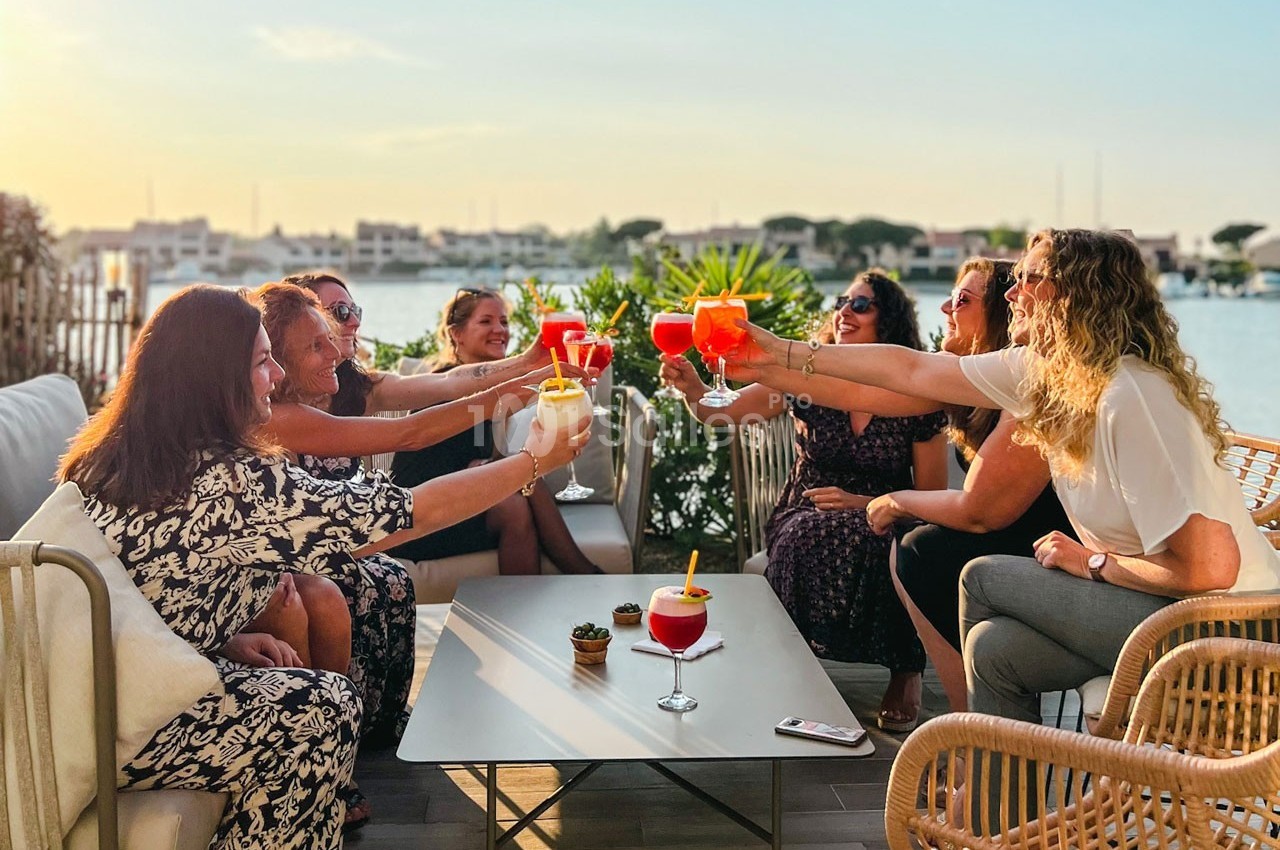 Un groupe de personnes trinque avec des cocktails en terrasse au bord de l'eau, sous une lumière de fin de journée.
