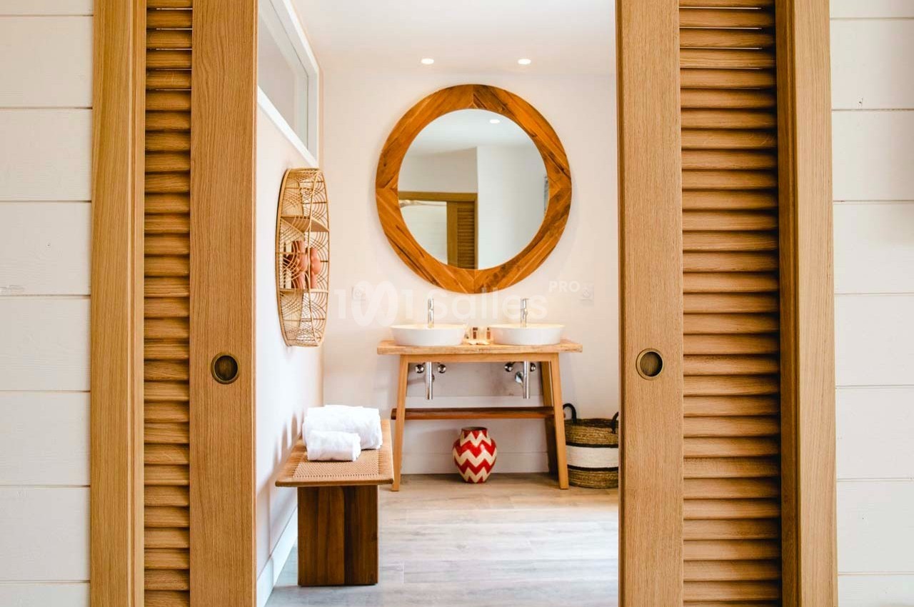 Salle de bain lumineuse avec deux vasques, miroir rond en bois, banc avec serviettes et éléments décoratifs.