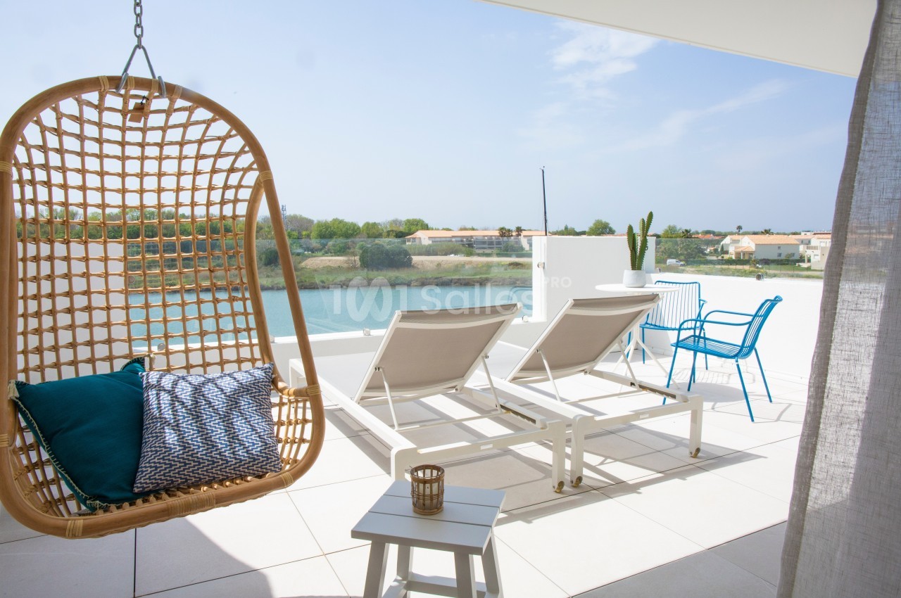 Terrasse avec vue sur un plan d'eau, équipée de transats, une chaise suspendue et une table avec des chaises.