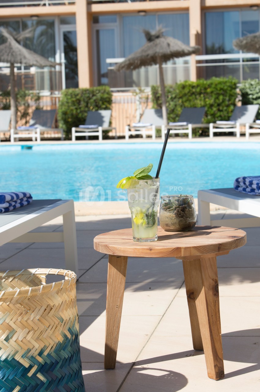 Verre de cocktail et plante sur une table en bois près d'une piscine entourée de transats et parasols.