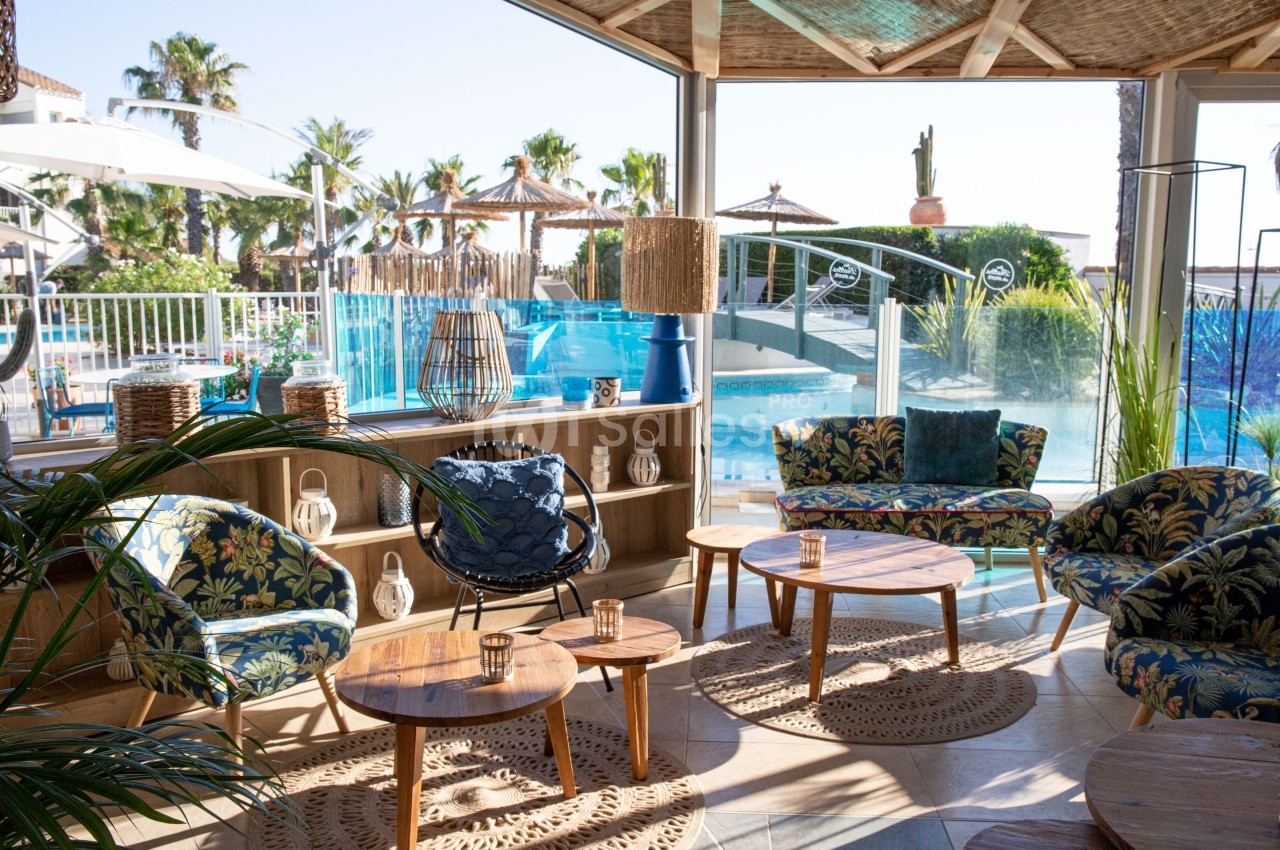 Espace lounge lumineux avec mobilier en bois et vue sur une piscine entourée de palmiers et parasols en paille.
