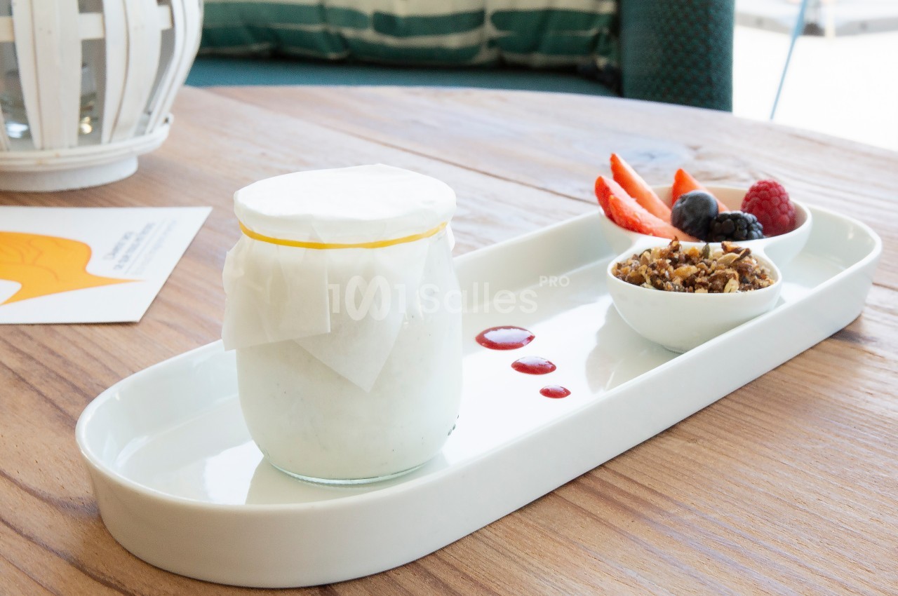 Pot de yaourt couvert de papier, accompagné de fruits rouges, granola et coulis sur un plateau blanc.