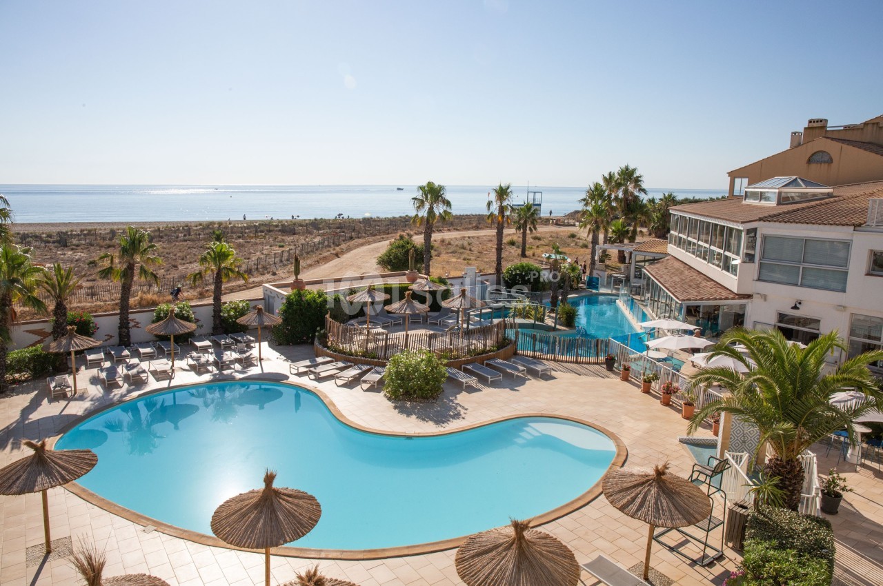 Vue d'une piscine extérieure avec transats, parasols en paille, palmiers et aperçu de la mer en arrière-plan.