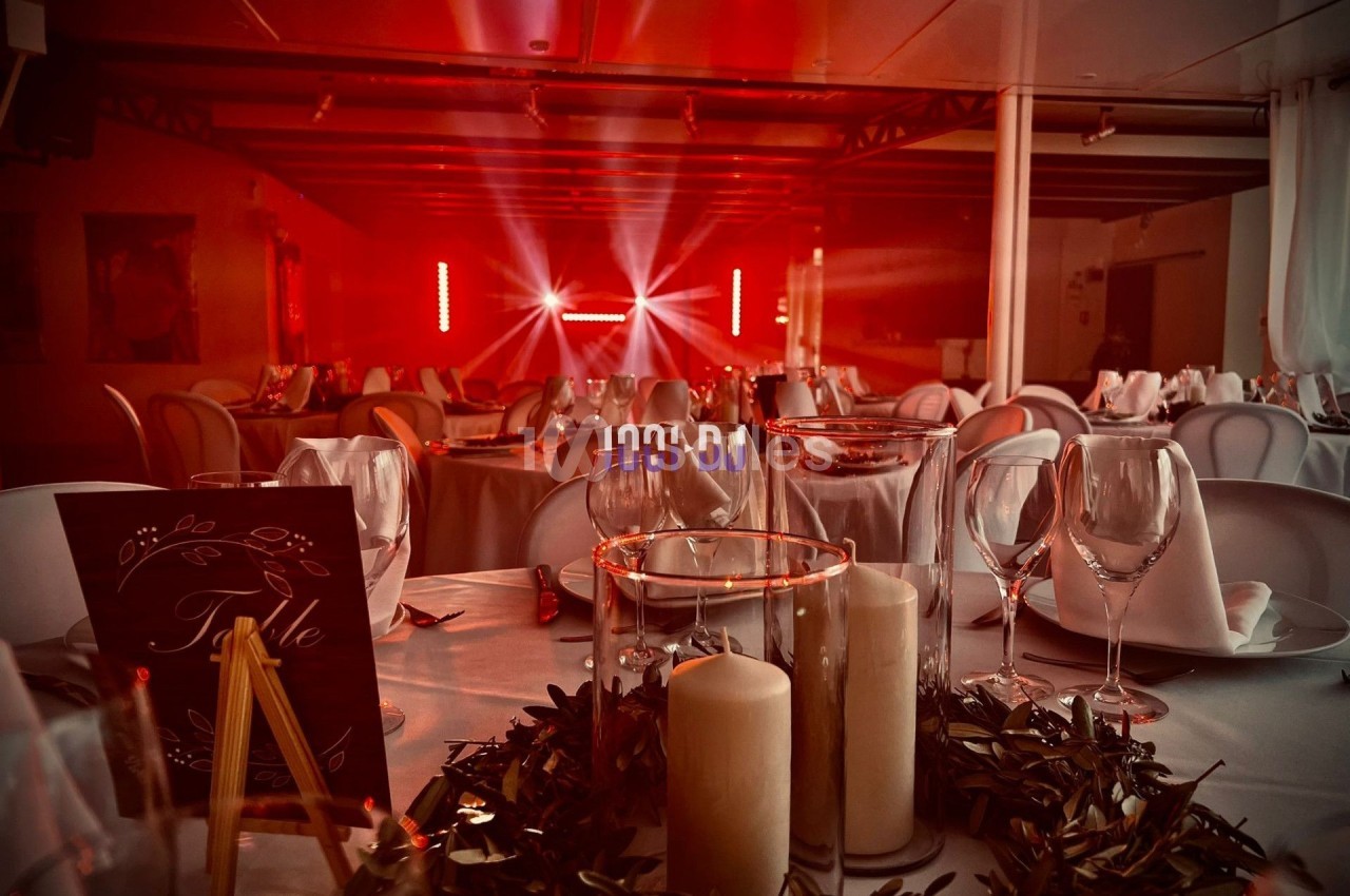 Salle de réception décorée avec des tables dressées, bougies et guirlandes, éclairée par des lumières rouges.