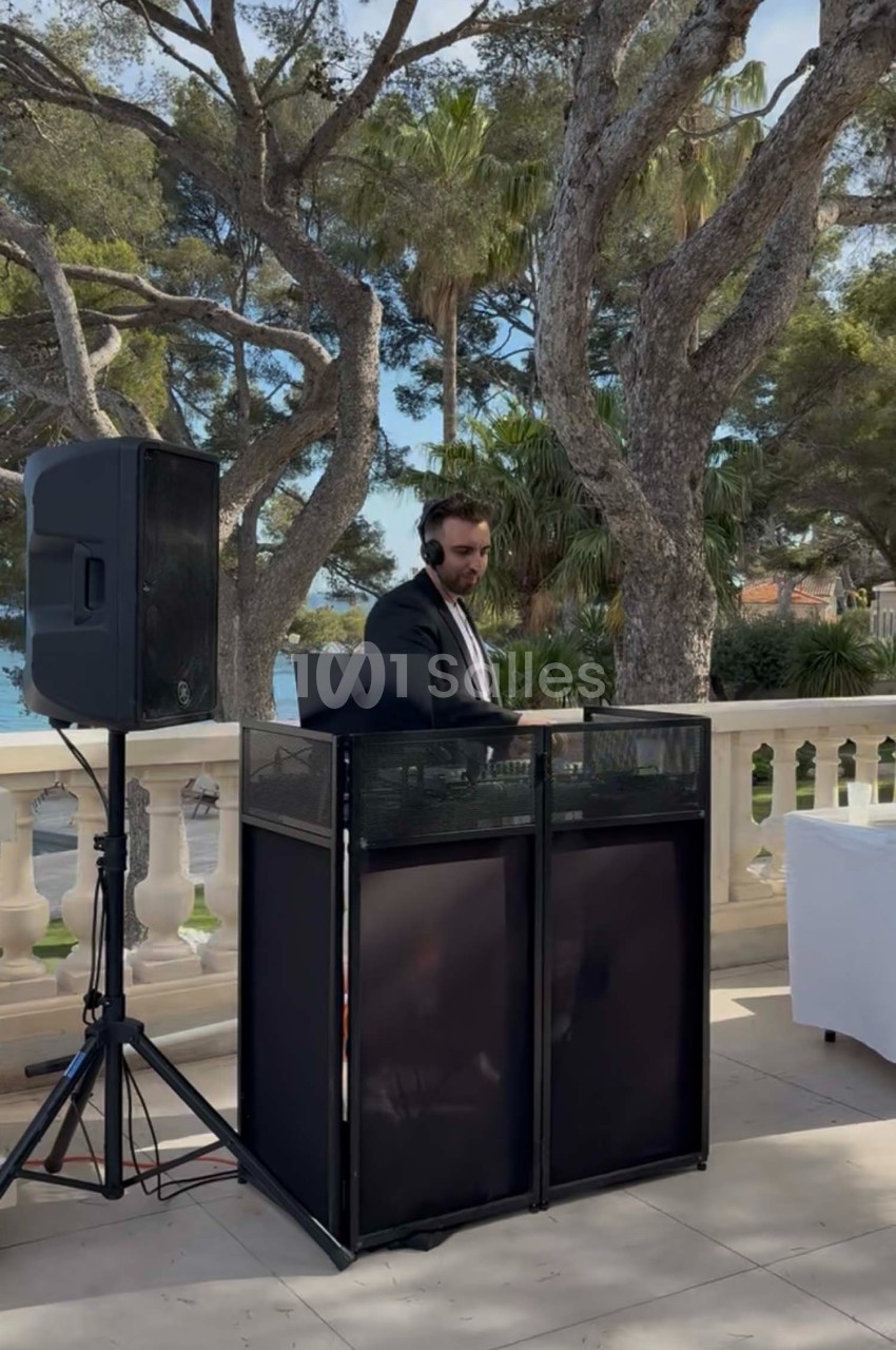 Un DJ en costume mixe de la musique en extérieur, devant une balustrade avec vue sur des arbres et la mer.