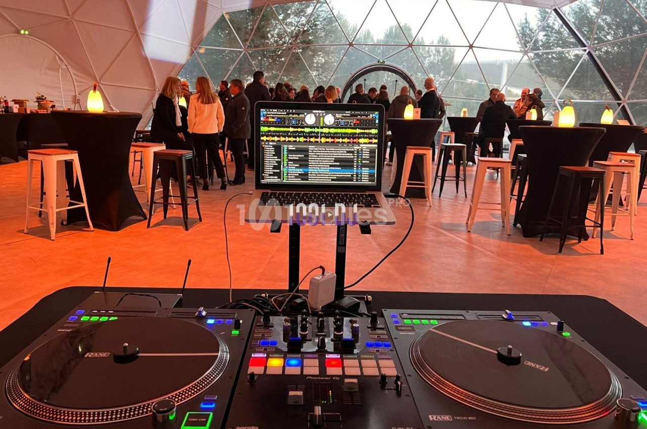 Console de DJ avec platines et ordinateur portable, devant un groupe de personnes réunies dans un espace sous une structure…